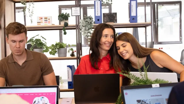 Zwei Frauen sitzen zusammen an einem Schreibtisch, eine in einem roten Hemd und die andere in einem grauen, und teilen einen Moment über Social-Media-Tools. Ein Mann in einem braunen Hemd arbeitet an einem separaten Schreibtisch. Im Hintergrund sind verschiedene Laptops und Pflanzen zu sehen.