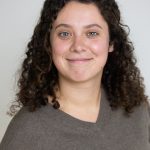 Eine junge Frau mit lockigem Haar lächelt vor weißem Hintergrund.