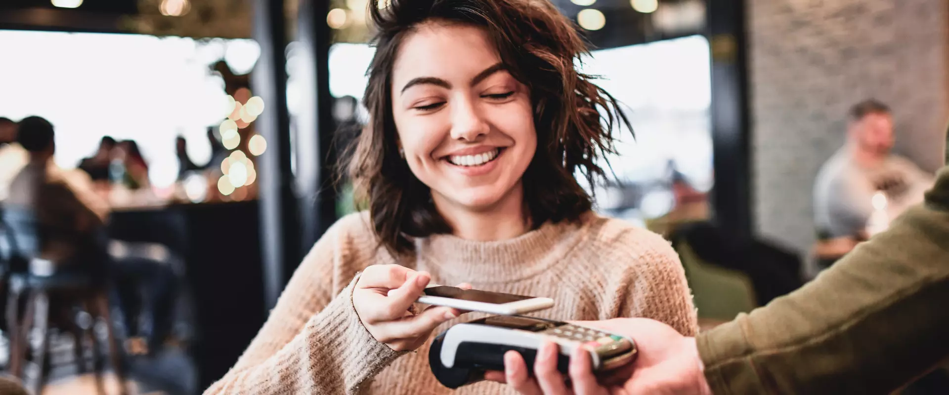 Eine lächelnde Frau benutzt ihr Smartphone, um in einem belebten Café an einem Kartenleser kontaktlos zu bezahlen.