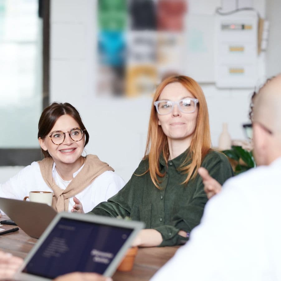 Zwei Personen sitzen an einem Tisch und unterhalten sich. Eine hält eine Tasse in der Hand, auf dem Tisch liegen Laptops. Eine andere Person, deren Gesicht teilweise sichtbar ist, spricht mit ihnen.
