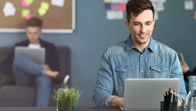 Ein Mann im Jeanshemd verwendet in einer geschäftigen Büroumgebung einen Laptop zur Marketingunterstützung, während im Hintergrund Kollegen arbeiten.