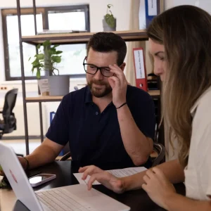 Zwei Mitglieder des Einfachmarketing-Teams arbeiten gemeinsam an einem Schreibtisch, umgeben von einem Laptop und Dokumenten. Der Mann rückt seine Brille zurecht, während die Frau auf dem Laptop tippt. Im Hintergrund sorgen Regale voller Ordner und Pflanzen für einen Hauch von Grün in ihrem produktiven Arbeitsplatz.
