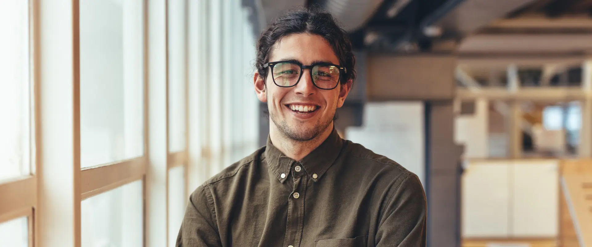 Ein Mann mit langen dunklen Haaren und Brille, der ein zugeknöpftes olivgrünes Hemd trägt, steht in einem Innenraum an großen Fenstern und lächelt in die Kamera - er strahlt das selbstbewusste Auftreten eines fraktionierten CMO aus.