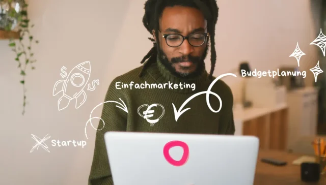 Ein Mann mit Brille arbeitet an einem Laptop, auf dem weiße marketingbezogene Kritzeleien und deutsche Wörter wie "Einfachmarketing", "Budgetplanung" und "Marketingkosten Startup" zu sehen sind.