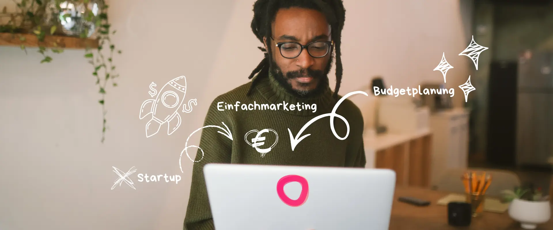Ein Mann mit Brille arbeitet an einem Laptop, auf dem weiße marketingbezogene Kritzeleien und deutsche Wörter wie "Einfachmarketing", "Budgetplanung" und "Marketingkosten Startup" zu sehen sind.