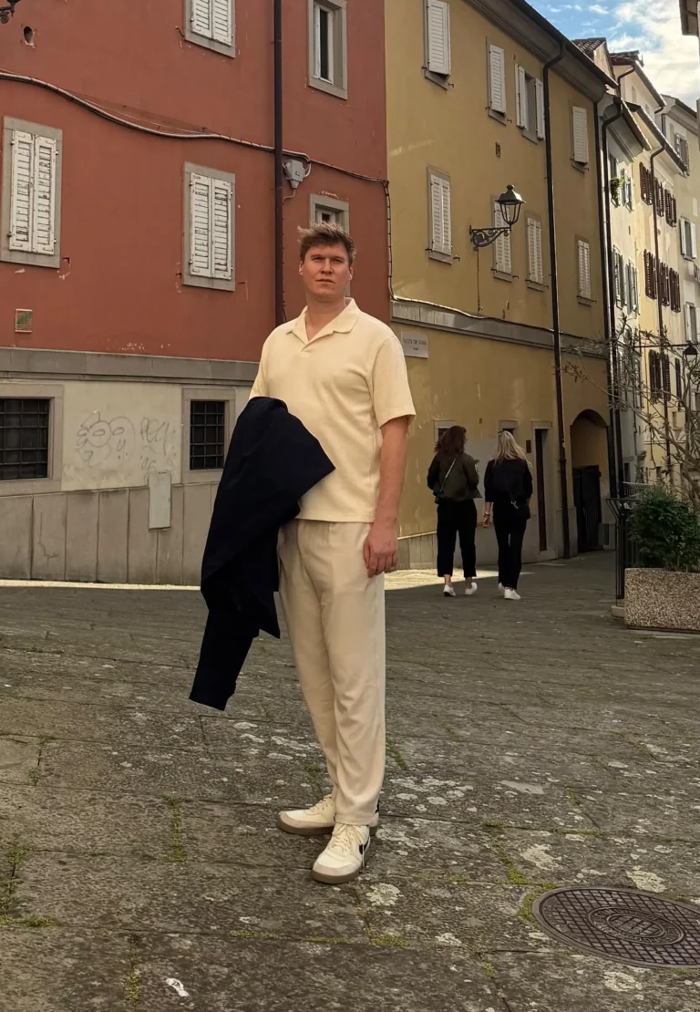 Leonhard Pleschberger in leichter Kleidung steht auf einem kopfsteingepflasterten Platz, umgeben von bunten Gebäuden unter einem bewölkten Himmel. Im Hintergrund sind ein paar Menschen zu sehen.