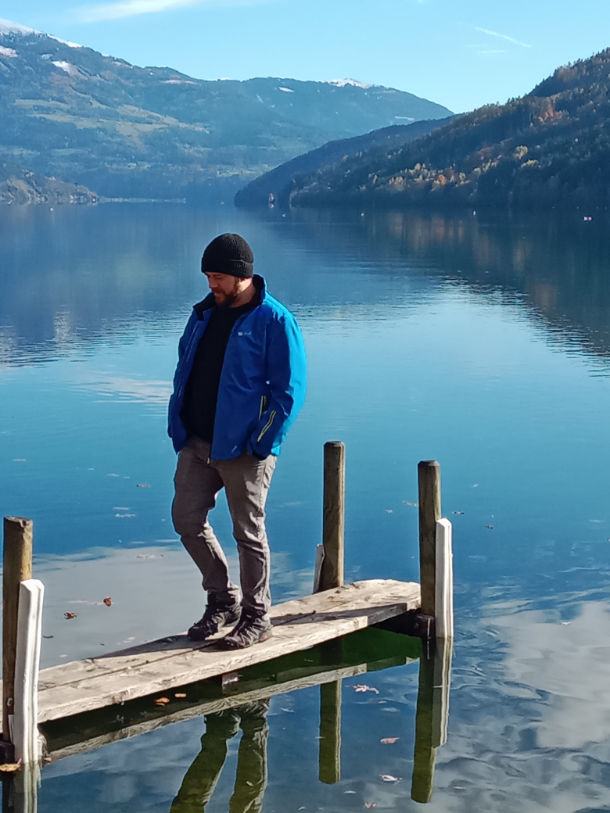 Leonhard Pleschberger, in blauer Jacke und schwarzer Mütze, steht auf einem Holzsteg mit Blick auf einen ruhigen See, der von Bergen und Bäumen eingerahmt ist.
