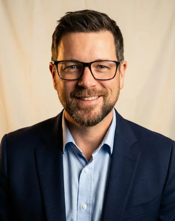 Mario Bernegger, ein Mann mit kurzen braunen Haaren, Bart und Brille, trägt eine marineblaue Anzugsjacke und ein hellblaues Hemd und lächelt vor einem schlichten beigen Hintergrund.