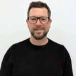 Vor einem schlichten weißen Hintergrund steht ein Mann mit kurzen Haaren, Bart und Brille, der einen schwarzen Pullover trägt und in die Kamera lächelt - ein freundliches Mitglied des Einfachmarketing-Teams.
